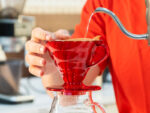 Artesano vertiendo agua caliente en un filtro de café de vidrio rojo, preparando café colombiano de origen natural en cafetería Raizal.